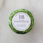 A round wrapped shower steamer from Hyssop Beauty Apothecary, featuring a shiny metallic green foil wrapper and a white circular label with black text.