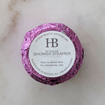 A round shower steamer wrapped in shiny purple foil with a white circular label reading 'Hyssop Beauty Apothecary HB All Natural Shower Steamer Lavender + Mint' on a light stone surface.