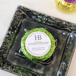 A round shower steamer wrapped in shiny green foil with a white circular label reading 'Hyssop Beauty Apothecary HB Shower Steamer,' placed on a dark decorative tray.